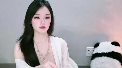 Video of a young Asian woman with long black hair and pale skin, wearing a white robe and necklace, seated beside a stuffed panda in a minimalistic, gray-toned room.