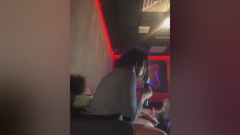 Video of a dimly lit, narrow hallway with red LED strip lighting, showing a person with dreadlocks in a grey shirt, holding a phone, and another person with curly hair lying on the floor.