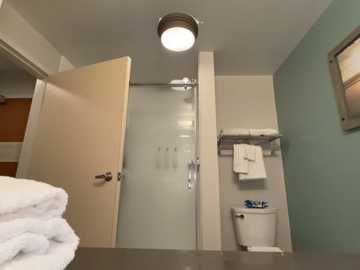 Video of a small, modern bathroom with a frosted glass door, beige walls, a white toilet, and neatly folded towels on a silver rack.