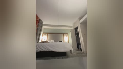 Media: Video of a modern, minimalist hotel room with white walls, beige carpet, and a large bed with white linens. The room features a light green accent wall, a tall window with white curtains, and a flat-screen TV on the wall.