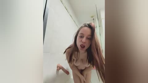Media: Video of a nude young woman with fair skin and long brown hair, bending over in a shower, partially obscured by steam, in a white-tiled bathroom.