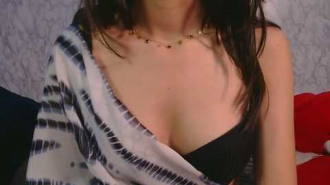 Video of a woman with fair skin, wearing a black bra and a tie-dye shirt, necklace, and long dark hair. Background features a textured wall and a red pillow.