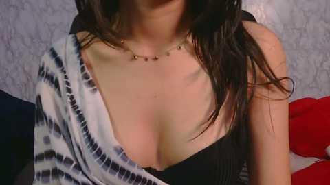 Video of a woman with long dark hair, wearing a black top with a plunging neckline, revealing ample cleavage. She has a thin gold necklace. Background features a textured grey wall and red and white decorative pillows.