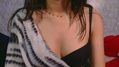 Video of a woman with wet, dark hair, wearing a black bra and a blue tie-dye shirt, sitting indoors with a patterned wallpaper background.
