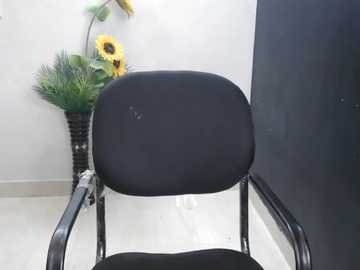 Media: Video of a black, cushioned office chair with metal arms, placed in a minimalist room with white walls and a vase of fake sunflowers and greenery on the left.