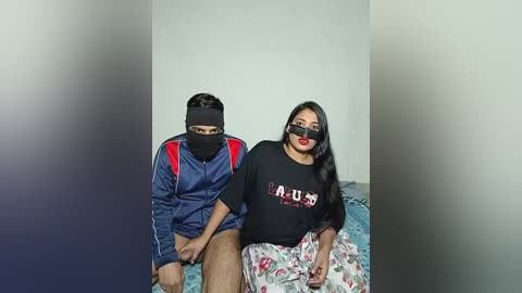 Video of two South Asian individuals seated on a bed. The man on the left wears a blue jacket, black mask, and beige shorts. The woman on the right has long black hair, black glasses, and red lipstick, wearing a black shirt with white flowers.