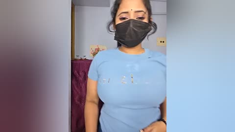 Video of a South Asian woman with medium skin tone and medium-length black hair, wearing a light blue short-sleeved shirt, black face mask, and a black headband, standing in a room with a purple curtain and a light-colored wall.