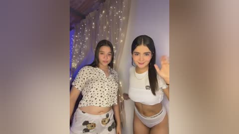Video of two young Latina women with medium brown skin and long black hair, one in a white polka-dot top and shorts, the other in a cropped white top and white panties, standing side by side in a dimly lit room with string lights and a curtain background.