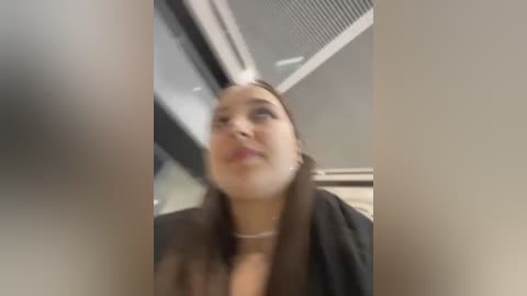 Video of a woman with medium skin tone, brown hair, and a black jacket, looking up with a neutral expression. Background shows a ceiling with air vents and fluorescent lights.