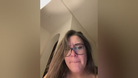 Video of a middle-aged woman with long, light brown hair, wearing black-framed glasses, looking slightly annoyed. She is indoors, in a room with arched doorways and white walls.