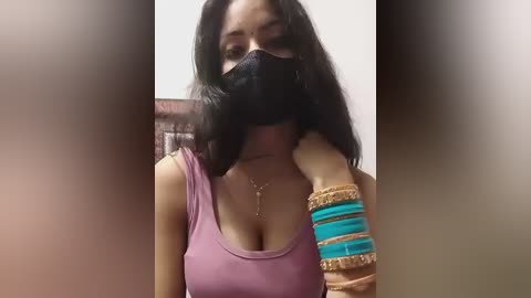 Media: Video of a South Asian woman with medium skin tone, wearing a black mask, purple tank top, and gold necklace, showing her cleavage, with arm bracelets, indoors.