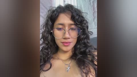 A video of a young woman with curly black hair, fair skin, and round glasses, wearing a silver necklace. She is topless, set against a blurred background with a white wall.
