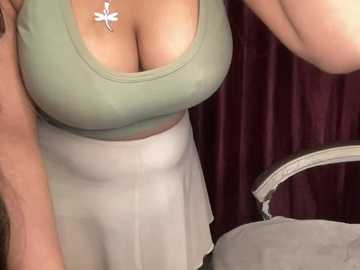 Media: Video of a woman with fair skin, large breasts, wearing a tight green tank top and white skirt, standing against maroon curtains. A silver cross necklace is visible.