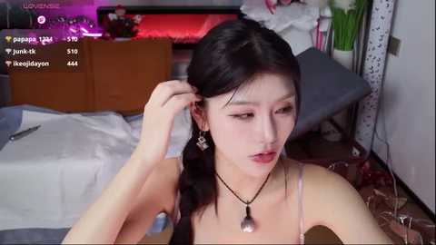 Video of an Asian woman with long black hair, wearing a spaghetti strap top, adjusting her hair in a bedroom with a bed, a table, and a TV.