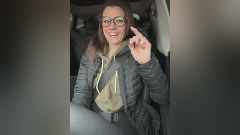 A video of a smiling, middle-aged woman with glasses, wearing a grey puffer jacket, olive-green hoodie, and grey pants, sitting in a car with a safety belt on.