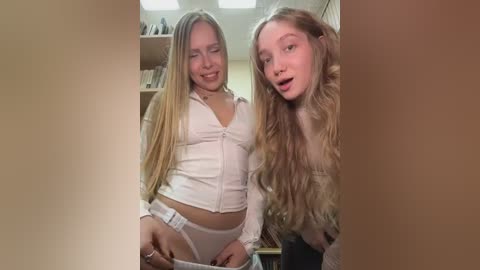 Video of two young women with long blonde hair, wearing matching white crop tops and white panties, standing in a book-filled room.