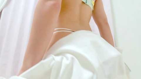 A video of a woman in a white bikini, partially obscured by a white sheet. Her back is turned, showing her tan lines and the straps of her bikini. The background is a plain white wall.
