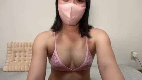 Media: A video of an Asian woman with medium skin tone and shoulder-length black hair, wearing a pink lace bra and face mask. She is seated in a minimalistic bedroom with beige walls and a tufted headboard.