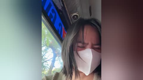 Media: Video of a young woman with straight, shoulder-length black hair, wearing a white mask and green shirt, seated in a car with tinted windows and a visible tree outside.