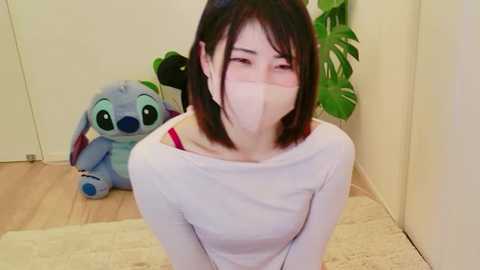 Video of an Asian woman with straight black hair, wearing a white long-sleeve top, beige face mask, and a red bra, kneeling on a beige rug. A blue Stitch plush toy and a green plant are in the background.