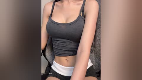 Video of a light-skinned woman with long, dark hair, wearing a tight, gray tank top and black shorts, sitting on a gray chair, indoor setting.