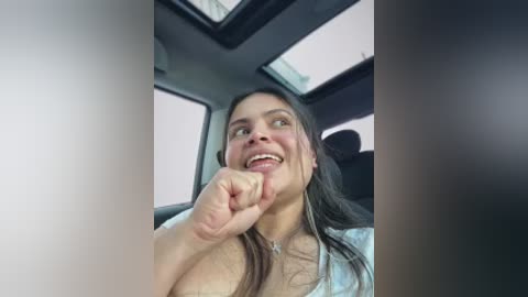 Video of a young woman with long dark hair, smiling widely, sitting in a car with a sunroof open, wearing a light-colored top.