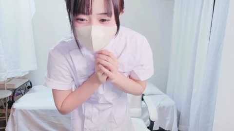 Video of a young East Asian woman in a white surgical mask, holding a white cloth to her face, standing in a clinical room with white curtains and medical equipment.