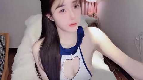 A video of an Asian woman with long black hair, fair skin, and a slender build, wearing a revealing white halter top with a blue bow, in a cozy, dimly-lit bedroom.