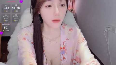 Video of an East Asian woman with fair skin, wearing a pink floral kimono, sitting on a white fur chair, in a bedroom with a gray wall. The image includes a live stream overlay with chat messages.