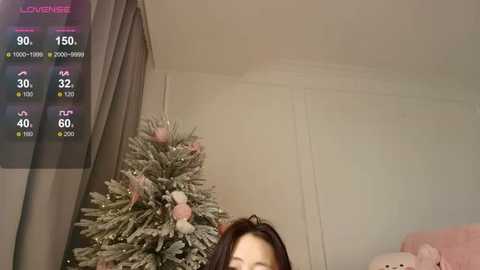 A video of a young woman with long dark hair partially visible, standing in a cozy room with a Christmas tree adorned with pink and white ornaments. The room has beige walls and a pink couch in the background.