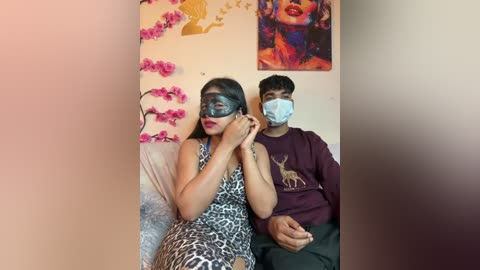Media: Video of a woman in a leopard dress and mask, sitting next to a man in a maroon sweatshirt with a deer graphic, both wearing face masks in a room with pink flowers and a colorful painting on the wall.