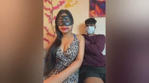 Video of a woman in a leopard-print dress with a black mask, seated in front of a man in a purple shirt, both indoors, with colorful wall art in the background.