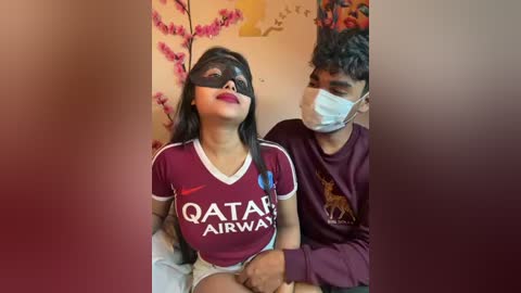 Video of a woman in a dark maroon Qatar Airways t-shirt, sitting with a man wearing a maroon shirt and a face mask, both indoors.