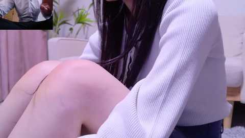 Video of a woman with long, dark hair in a white ribbed sweater, leaning forward, revealing her bare buttocks. Background shows a white couch, green plant, and a TV.