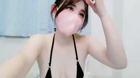Video of a young Asian woman with fair skin, wearing a black bikini and a pink face mask, adjusting her hair in a white-tiled bathroom.