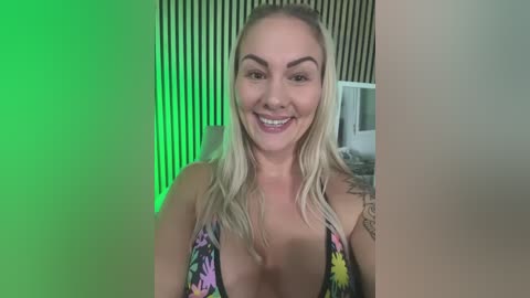 Video of a smiling, fair-skinned woman with long blonde hair, wearing a colorful, low-cut bikini top, standing against a green-lit, striped wall, with a tattoo on her upper arm.