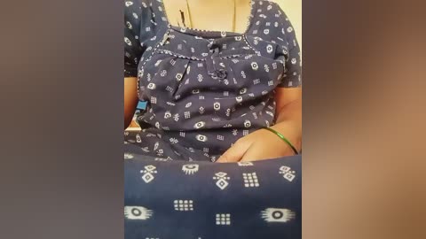 Video of a person wearing a blue, short-sleeved, patterned nightgown with white geometric designs. The background is blurred, showcasing a beige-colored surface. The person's arms are crossed, and a green bracelet is visible on their wrist.