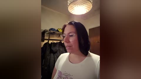 Video of a woman with shoulder-length dark hair in a white t-shirt, standing in a dimly lit room with a chandelier. Behind her, a rack holds black jackets and a yellow helmet.