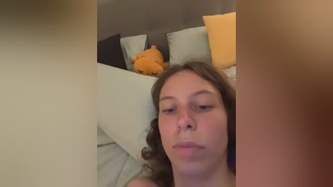 A video of a young woman with long brown hair, lying on a bed with a plush toy and multiple pillows in a cozy, dimly lit bedroom.