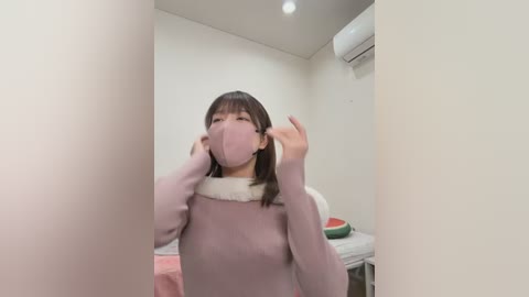 Video of an Asian woman with straight black hair and fair skin, wearing a pink face mask and a light purple sweater with white fur collar, in a simple, white-walled room with a small table and a watermelon on it.