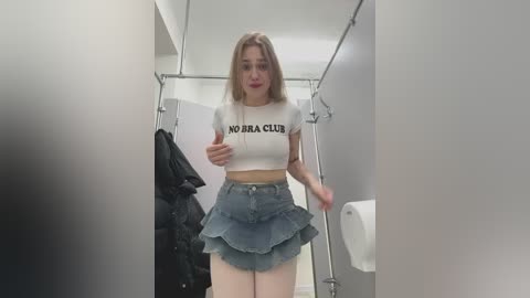 A video of a young woman with light skin, long blonde hair, and a slender physique, wearing a white crop top and high-waisted denim skirt, in a public restroom.