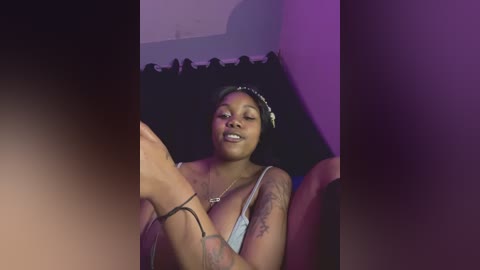 A video of a young Black woman with a medium-dark complexion, wearing a white bra and black hairband, sitting in a chair with a purple backdrop. She has a tattoo on her left forearm.