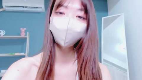 Video of an East Asian woman with long, straight brown hair, wearing a white surgical mask, in a room with blue walls, shelves, and an air conditioning unit.