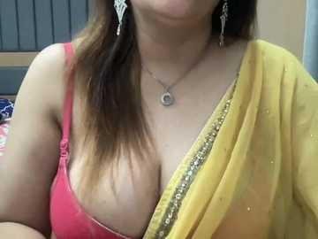 Video of a woman in a yellow sari with a red blouse, showcasing her cleavage, wearing a silver pendant necklace and dangling earrings. The background shows a beige wall and a window.
