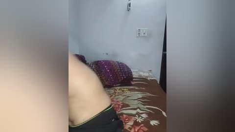 Media: Video of a small, dimly lit bedroom with a purple patterned cushion on a floral-patterned bedspread, a white wall with a power outlet, and a blurred figure partially visible in the foreground.