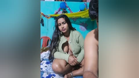 Video of a young South Asian woman with long black hair, wearing a green sweater, sitting cross-legged on a blue blanket. Background features a teal wall decorated with colorful streamers.