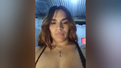 Video of a Latina woman with medium skin tone, shoulder-length wavy brown hair, and wearing a black spaghetti-strap top. She has a neutral expression and is indoors with a corrugated metal ceiling and exposed concrete walls.