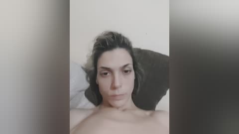 A video of a shirtless, light-skinned man with short, dark hair lying on a bed with a white pillow. The image is partially blurred, focusing on his upper torso and face.