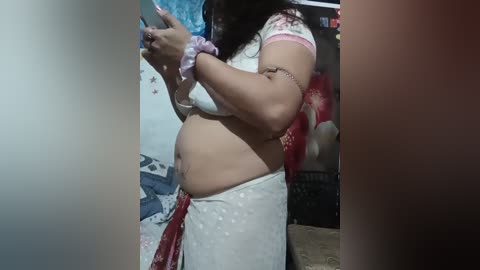 Video of a plus-sized woman in a white saree with a red border, showing her belly and wearing a pink scrunchie. Background includes a wall with blue and red decorations, and a bed with blue and white bedding.