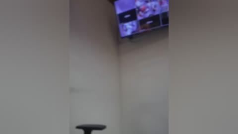 Blurry video of a beige room with a flat-screen TV displaying several small images. A black chair is partially visible in the lower foreground. The scene is dimly lit, with a slight purple hue.